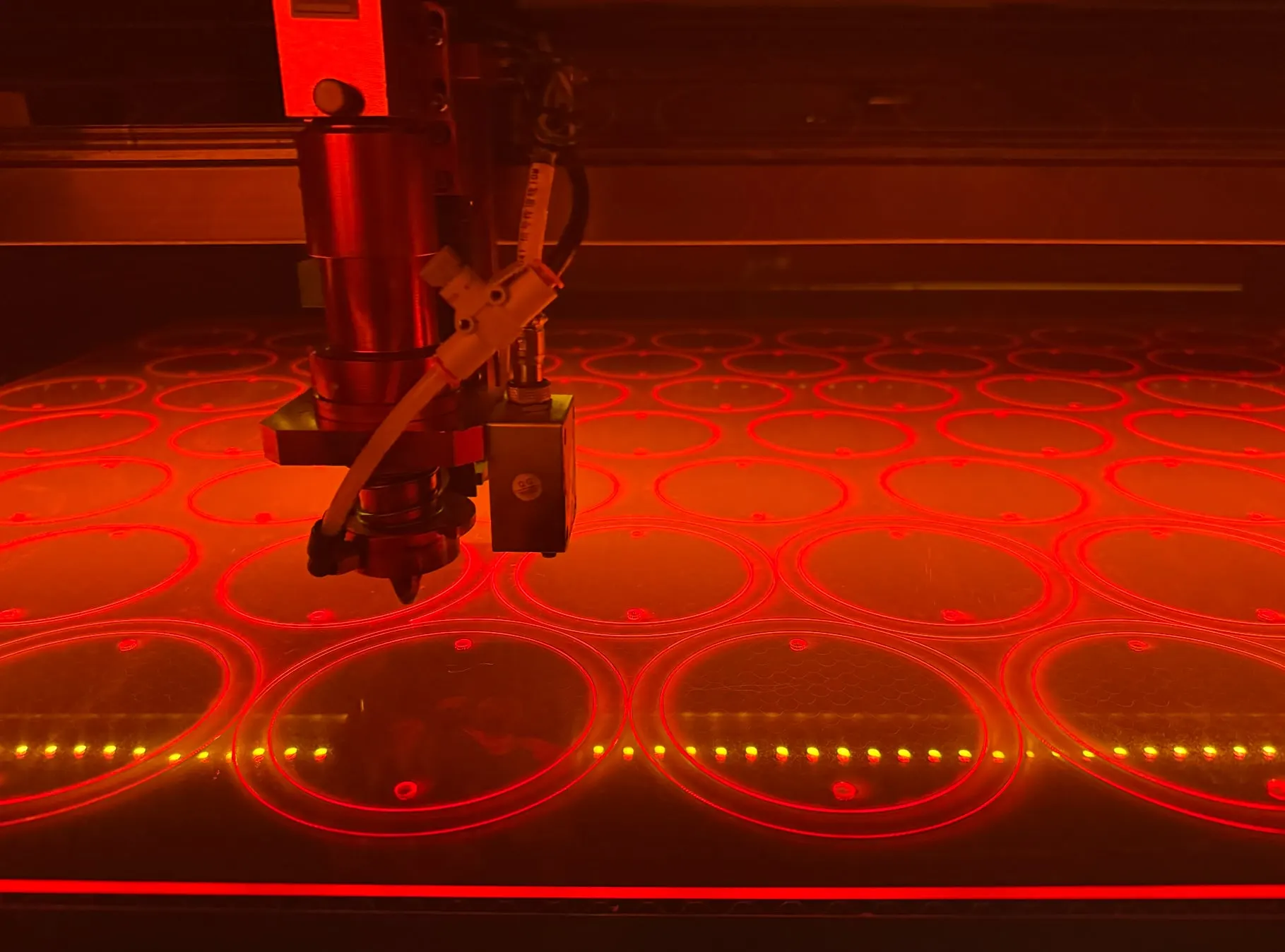 A CO2 laser head cutting rows of circles from a glowing red acrylic sheet
