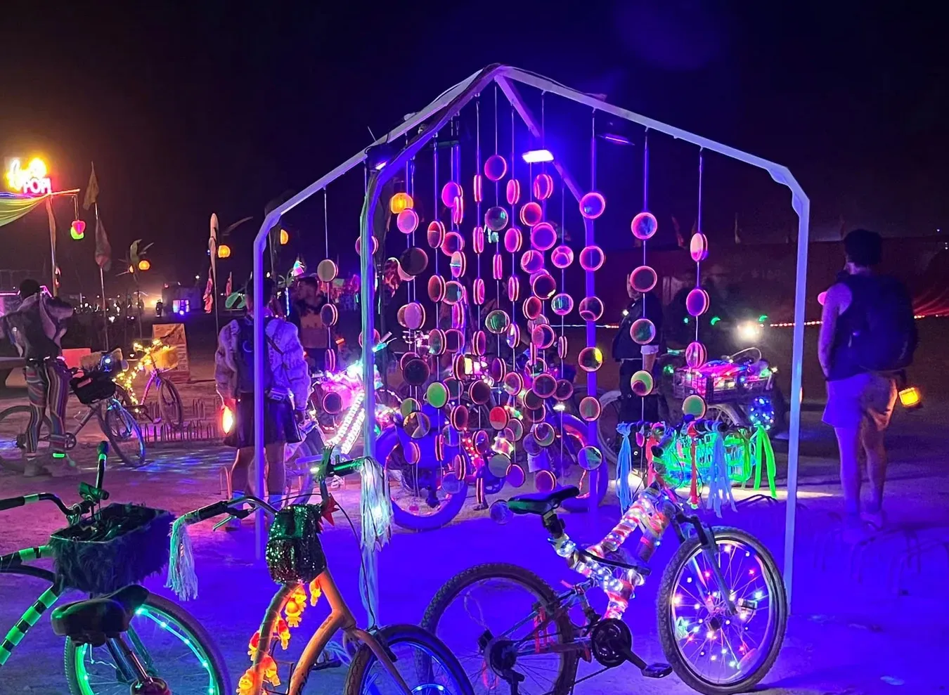 The installation at night on the playa, its frame and hanging mirrors glowing pink and violet under UV, with lit-up bikes parked in front